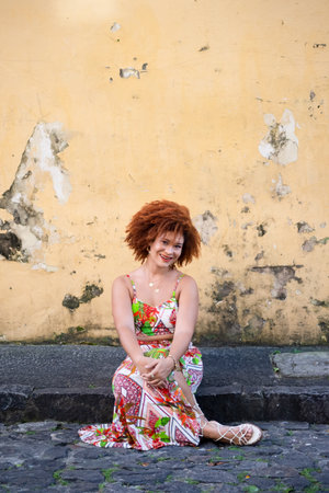 Portrait of a beautiful woman with red hair, in a long colorful dress, sitting on the sidewalk of a...