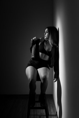 Beautiful young Caucasian woman sitting on a wooden stool, wearing casual clothes. Black and white portrait in the studio.の写真素材