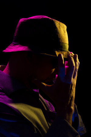 Portrait of a serious mysterious man with a beard wearing a hat and sunglasses making gestures with his hands. Portrait in studio with colored lights and dark background.の写真素材