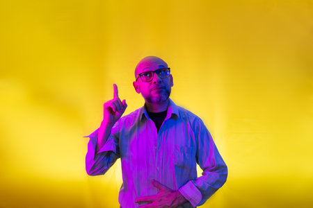 Bald, bearded man wearing glasses pointing up. Isolated on yellow background.