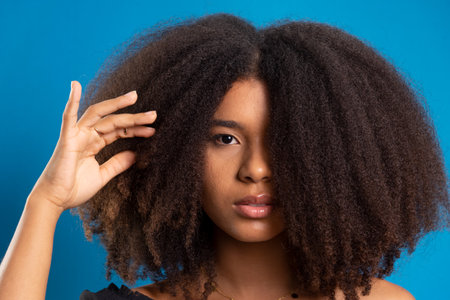 Half-closed portrait of beautiful young woman touching black power hair. Isolated on blue background.の写真素材