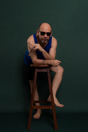 Bearded, bald white man wearing sunglasses leaning on wooden stool posing for photo. Isolated on dark green background.の写真素材