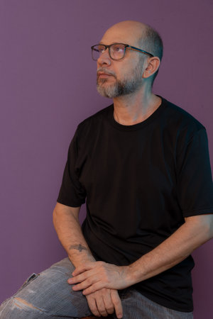 Portrait of bearded, bald man wearing prescription glasses sitting on wooden stool posing for photo. Isolated on dark lilac background.の写真素材