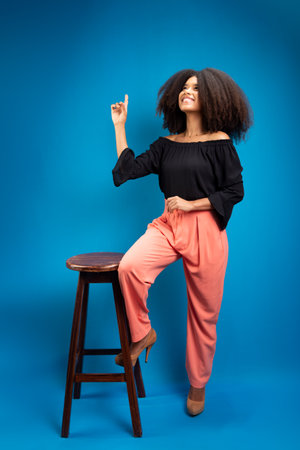 Young woman with natural curly hair, leaning on a wooden stool pointing upwards. Isolated on blue background.の写真素材