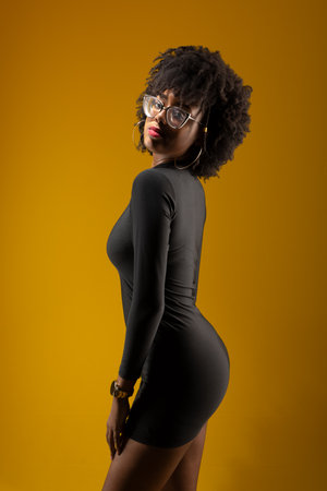 Beautiful sensual young woman wearing prescription glasses, standing, wearing black clothes against yellow background.の写真素材