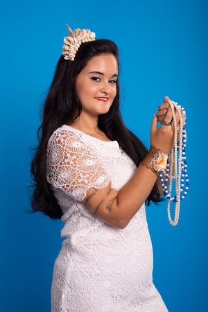 Beautiful Caucasian woman in a white dress with a shell tiara on her head holding pearl necklaces...