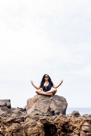 Beautiful, curly-haired woman sitting on the rock of a beach doing yoga exercise. Happy person traveling.の写真素材