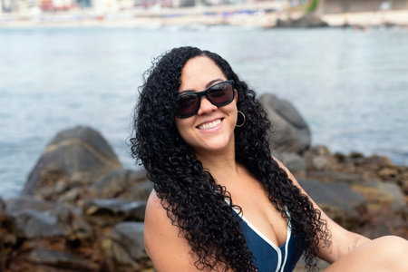 Beautiful, curly-haired woman wearing sunglasses looking at the camera against the sea in the background. Happy person traveling.の写真素材