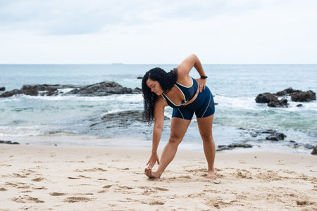 Beautiful, curly-haired, young woman in gym clothes doing stretches on the beach sand. Healthy lifestyle.の写真素材