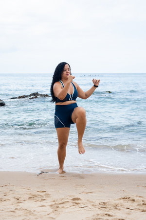 Fitness woman, standing, doing leg exercises facing the camera on the beach sand against the sea and cloudy sky. Healthy lifestyle.の写真素材