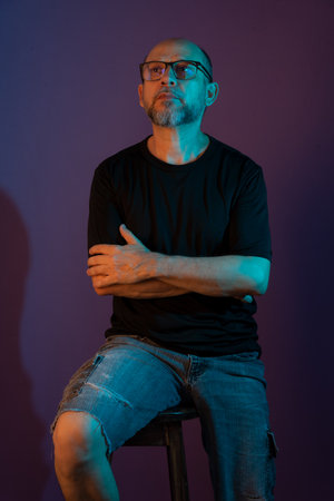 Portrait of bearded, bald man wearing prescription glasses sitting on wooden stool posing for photo. Isolated on dark lilac background.の写真素材