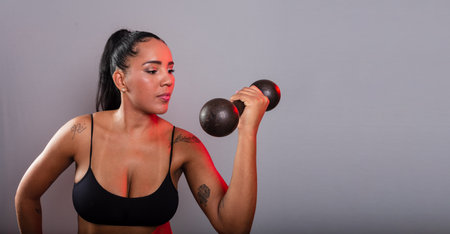 Beautiful brunette woman, in gym clothes, doing exercises with her arm holding black dumbbells. Healthy life. Against gray background.の写真素材