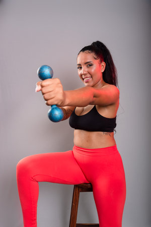 Beautiful, brunette, happy and smiling woman in gym clothes, holding dumbbells and looking at the camera. Healthy life. Against gray background.の写真素材