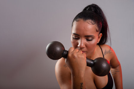Beautiful brunette woman, in gym clothes, sitting holding dumbbells at face level. Healthy life. Against gray background.の写真素材