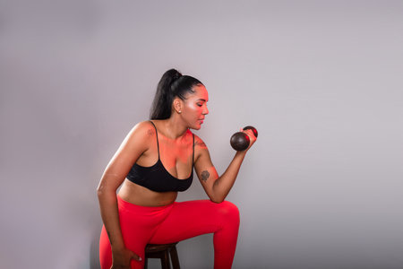 Beautiful brunette woman, in gym clothes, sitting doing exercises with dumbbells. Healthy life. Against gray background.の写真素材