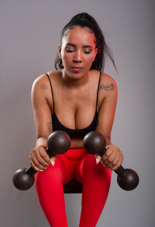 Beautiful brunette woman, in gym clothes, sitting facing the camera holding two dumbbells. Healthy life. Against gray background.の写真素材