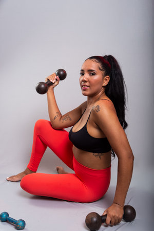 Athlete, brunette, happy and smiling woman in gym clothes, sitting doing exercises with dumbbells. Healthy life. Against gray background.の写真素材
