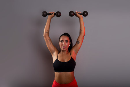 Beautiful brunette woman, in gym clothes, holding black dumbbells above her head. Healthy life. Against gray background.の写真素材