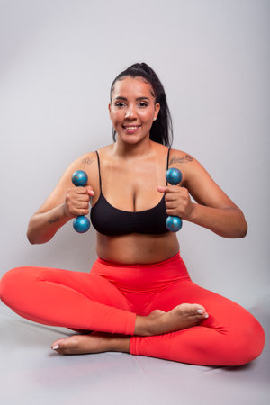 Beautiful, brunette, happy and smiling woman in gym clothes, sitting holding dumbbells. Healthy life. Against gray background.の写真素材