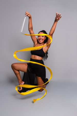 Gymnast woman, kneeling on the floor, doing movements with yellow ribbon. Isolated on gray background.の写真素材
