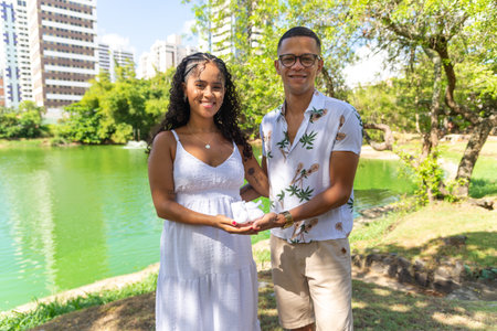 Married man and wife hold baby shoes in their hands. She is pregnant. Gender reveal. In the background a green lagoon.の写真素材