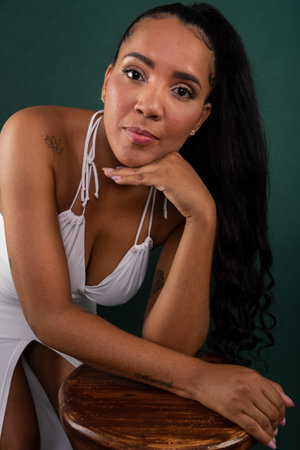 Beautiful brunette woman leaning on wooden stool with hand on chin posing for photo. Confident and happy person. Isolated on green background.の写真素材