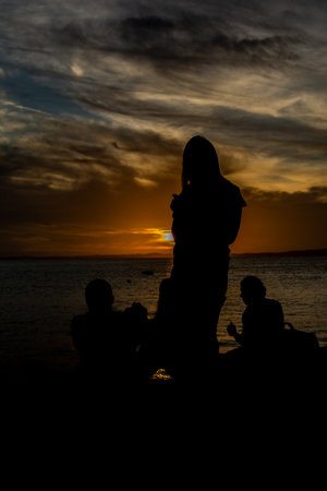 Silhouette of unidentified people enjoying dramatic sunset with dark clouds and sun in yellow color. City of Salvador, Bahia.の写真素材
