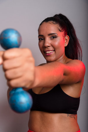 Beautiful, brunette, happy and smiling woman in gym clothes, holding dumbbells and looking at the camera. Healthy life. Against gray background.の写真素材