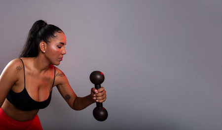 Beautiful brunette woman, in gym clothes, doing exercises with her arm holding black dumbbells. Healthy life. Against gray background.の写真素材