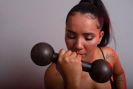 Beautiful brunette woman, in gym clothes, sitting holding dumbbells at face level. Healthy life. Against gray background.の写真素材