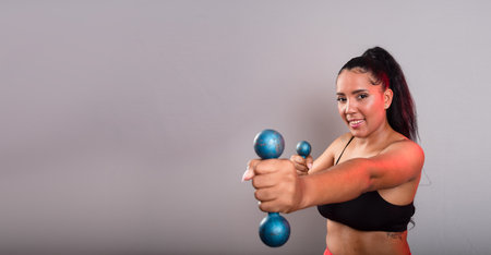 Beautiful, brunette, happy and smiling woman in gym clothes, holding dumbbells and looking at the camera. Healthy life. Against gray background.の写真素材