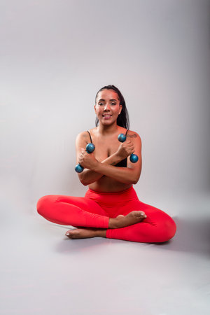 Beautiful, brunette, happy and smiling woman in gym clothes, sitting holding dumbbells with arms crossed looking at the camera. Healthy life. Against gray background.の写真素材