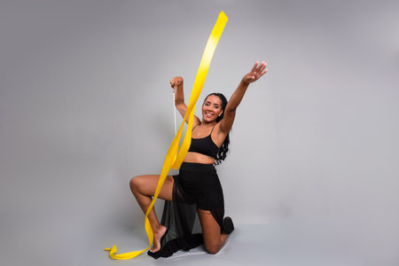 Gymnast woman, kneeling on the floor, doing movements with yellow ribbon. Isolated on gray background.の写真素材