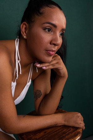 Beautiful brunette woman leaning on wooden stool with hand on chin posing for photo. Confident and happy person. Isolated on green background.の写真素材