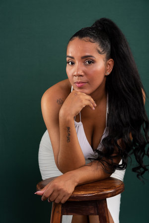 Beautiful brunette woman leaning on wooden stool with hand on chin posing for photo. Confident and happy person. Isolated on green background.の写真素材