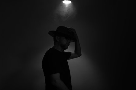 Black and white portrait of bearded man wearing hat with a light above. Studio portrait. Gray colored background with artificial smoke.の写真素材