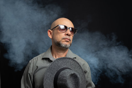 Man with a beard and sunglasses holding a gray hat. Against smoky background.の写真素材