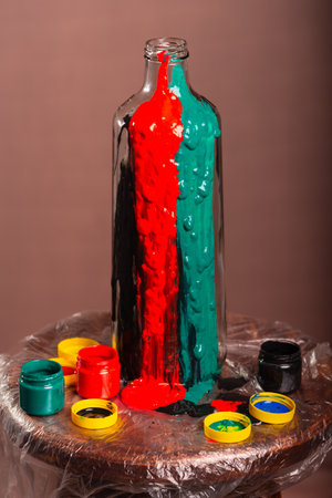 Closed portrait of a glass bottle with pots of paints of white, black, yellow, green, blue and red colors. Abstract. Isolated on brown background.の写真素材