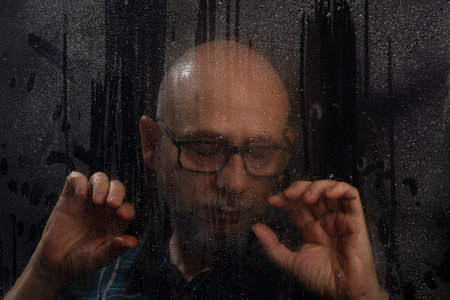 Close-up portrait of bearded bald man in prescription glasses touching wet glass. Isolated on black background.の写真素材