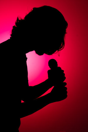 Silhouette of young man holding wireless microphone singing music with head down. Isolated on red background.の写真素材