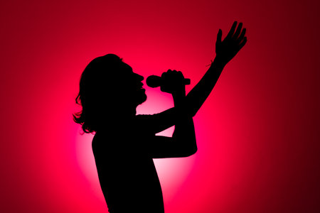 Silhouette of young man holding wireless microphone singing music and with his hand raised upwards. Isolated on red background.の写真素材