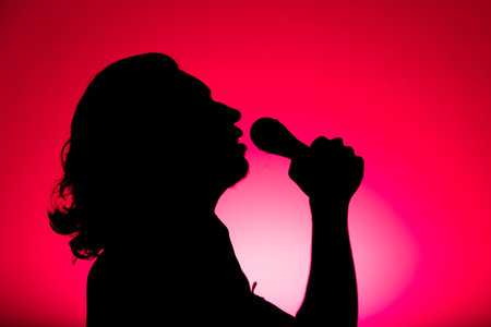 Silhouette of young man holding wireless microphone and singing music with emotion. Isolated on red background.の写真素材