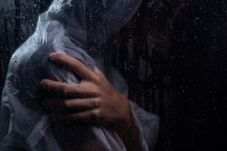 Close-up portrait of man wearing raincoat behind wet glass with hand holding right shoulder. Studio portrait. Isolated on dark background.の写真素材