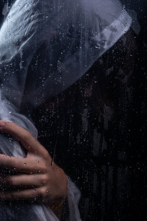 Close-up portrait of man wearing raincoat behind wet glass with hand holding right shoulder. Studio portrait. Isolated on dark background.の写真素材
