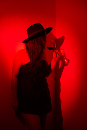 Portrait of beautiful woman in black clothes and hat drinking wine from a glass leaning against a wall. Studio photo with red gel.の写真素材