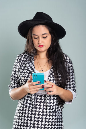 Young beautiful woman standing in studio looking at cell phone. Wearing black hat against light green background.の写真素材