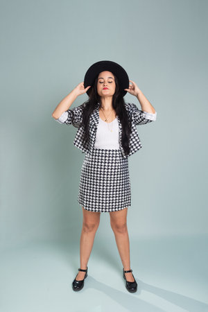 Young beautiful woman standing in studio posing for photo. Wearing and holding a black hat. Calm woman at ease wearing black and white clothing. Isolated on light green background.の写真素材