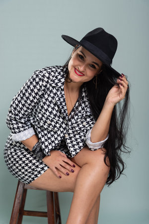 Beautiful smiling woman sitting with black hair wearing a black hat posing for photo in black and white outfit. Isolated on light green background.の写真素材