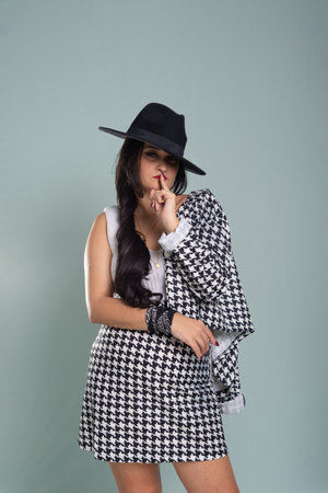 Portrait of a beautiful young woman standing wearing a black hat and black and white clothes making a sign with her finger on her mouth. Isolated on light green background.の写真素材