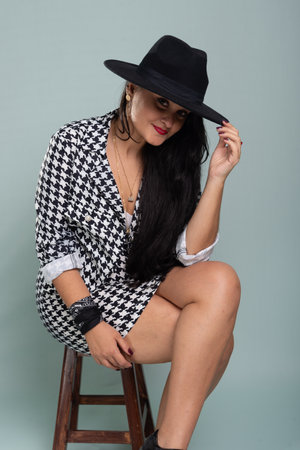 Beautiful smiling woman sitting with black hair wearing a black hat posing for photo in black and white outfit. Isolated on light green background.の写真素材
